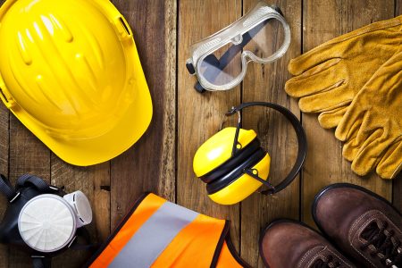 Personal safety workwear items shot directly above. The safety equipment includes a hard hat, goggles, gloves, steel toe shoes, earmuff a respiratory mask and a safety vest. The background is a rustic wood table scratched and stained with vertical stripes.  Predominant colors are yellow and brown. DSRL studio photo taken with Canon EOS 5D Mk II